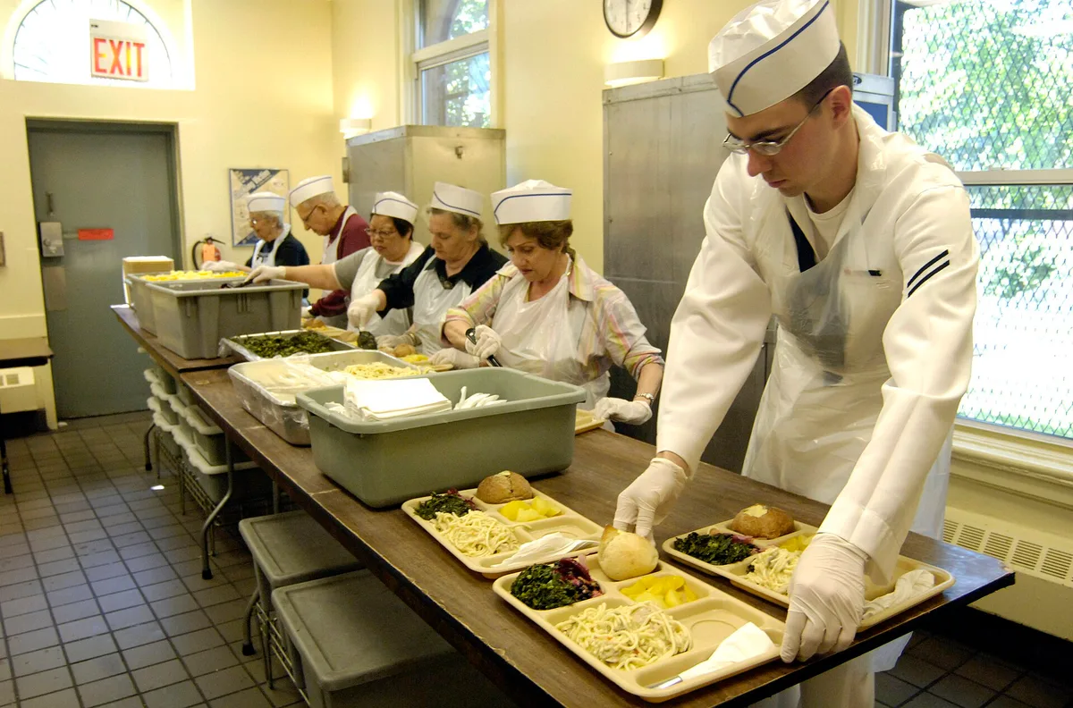 Voluntários preparam e servem bandejas de comida em uma cozinha comunitária que atende pessoas necessitadas