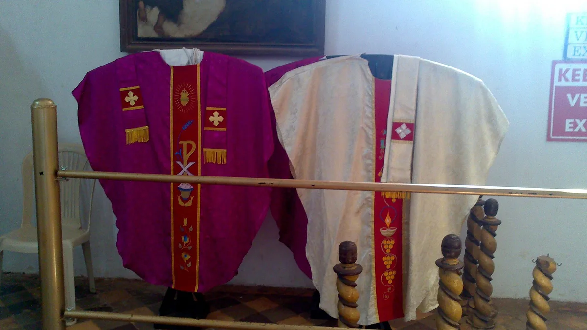Casulas litúrgicas roxa e branca expostas lado a lado em uma sacristia, mostrando cores diferentes dos paramentos
