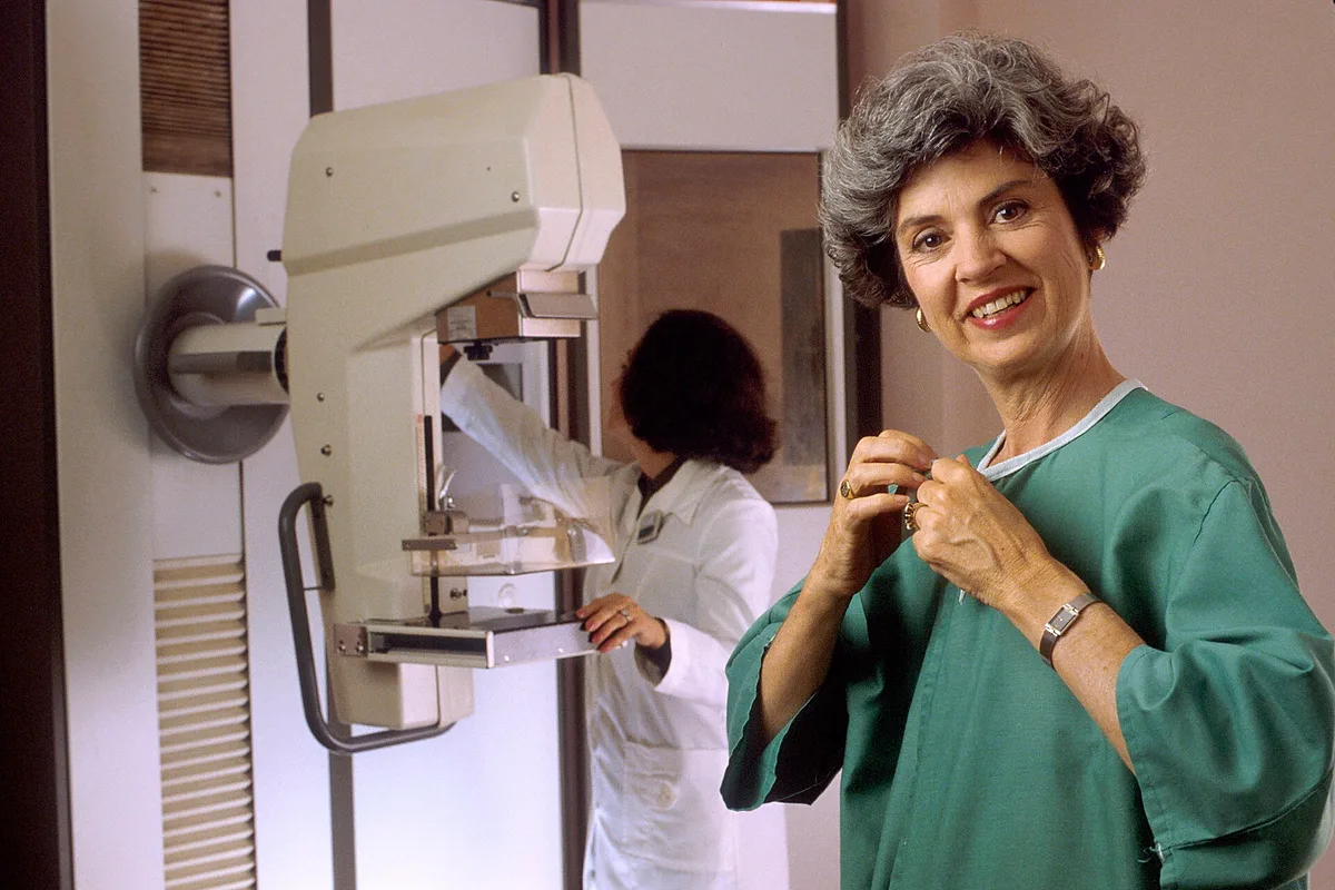 Mulher sorridente de avental hospitalar ao lado de um mamógrafo, com uma técnica ao fundo