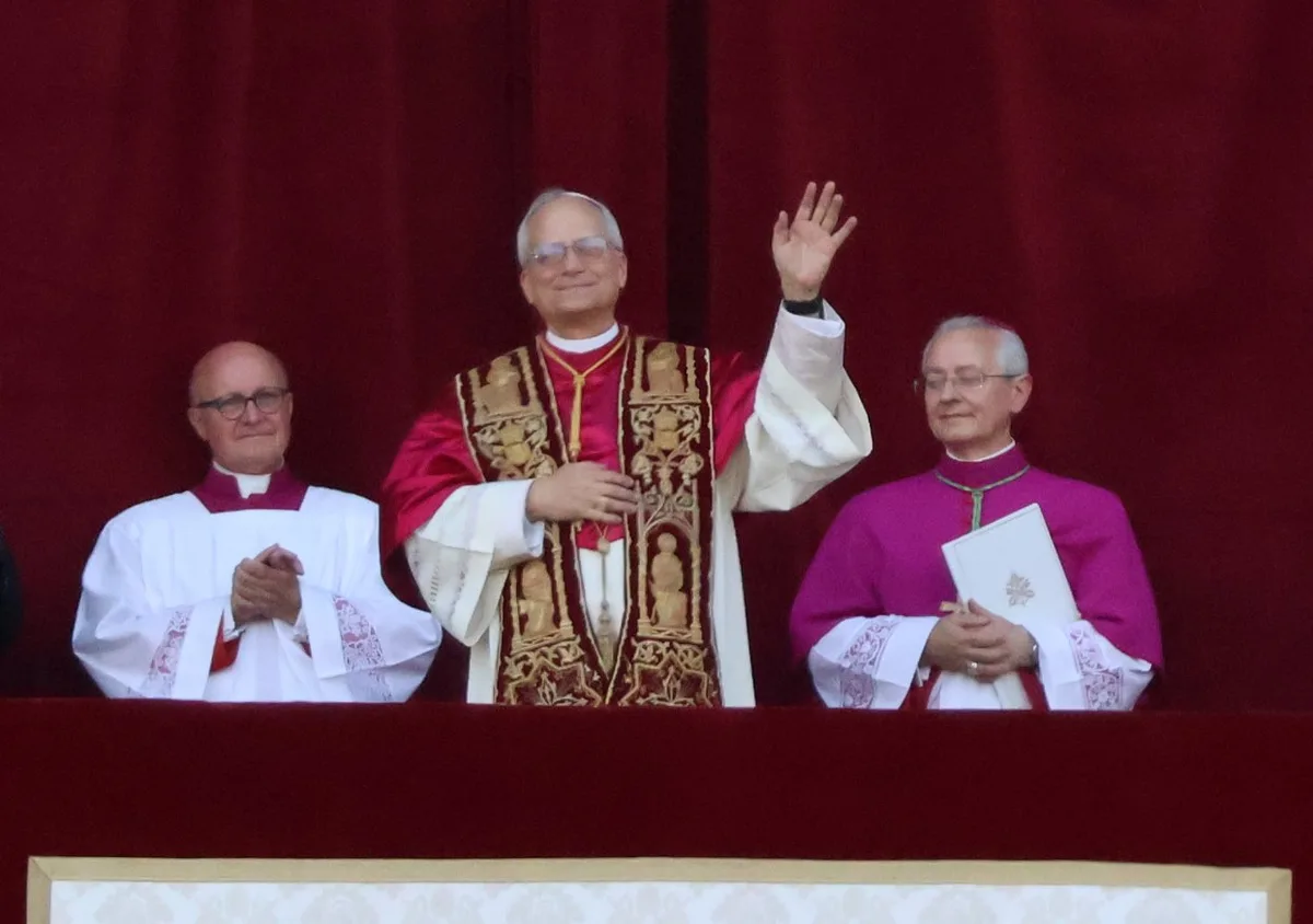 Papa Leão XIV acena da loggia, com vestes litúrgicas, ladeado por dois clérigos