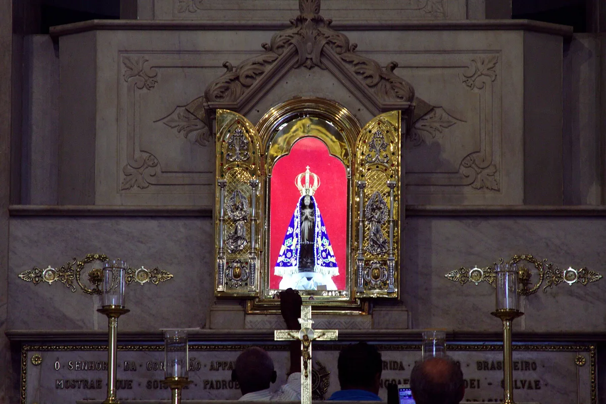 Imagem de Nossa Senhora Aparecida em oratório dourado com fundo vermelho, com fiéis rezando diante dela