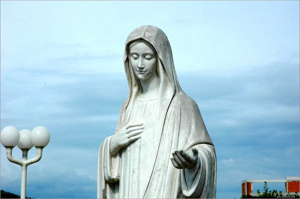 Estátua branca de Nossa Senhora de Medjugorje, com a mão no peito e a outra estendida, contra o céu azul