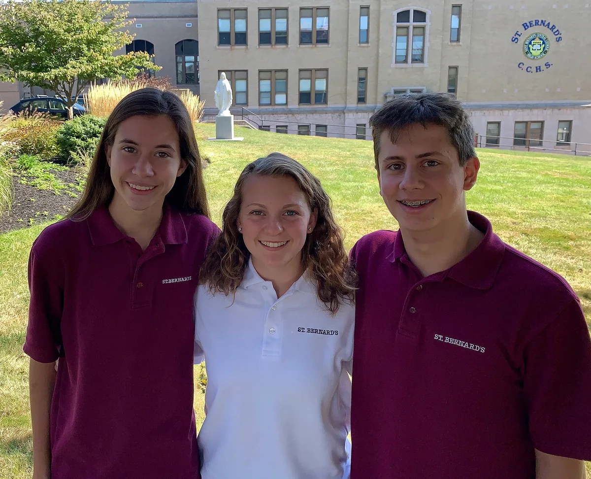 Três jovens estudantes sorridentes e abraçados, com uniforme de escola católica, no pátio gramado da escola