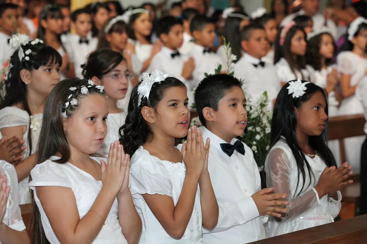 Crianças de branco, de mãos postas, rezando na igreja durante a Primeira Comunhão